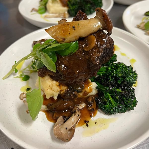 Braised short rib with mashed potatoes, broccolini, mushrooms, and gravy on a white plate.