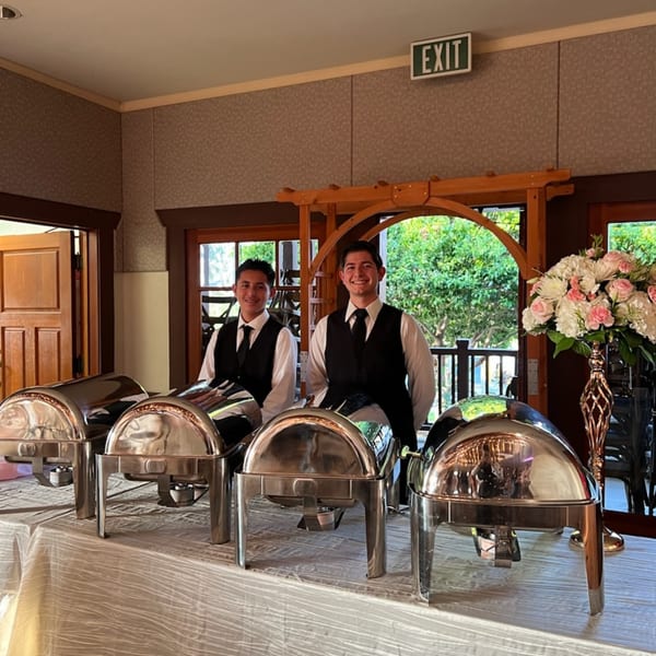 Two smiling Martiniz Fresko Kitchen staff stand behind a buffet table with chafing dishes.