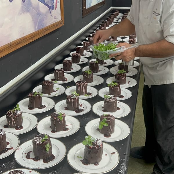 A chef garnishes rows of individual chocolate cakes with fresh greens on a long black table.