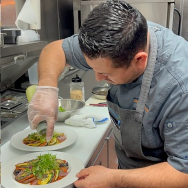 Chef in a gray shirt and apron garnishing two plates of colorful vegetable dishes in a kitchen.