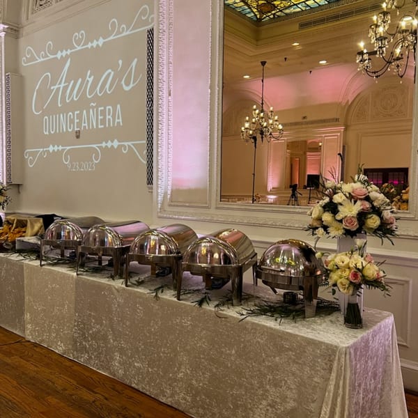 A buffet table with chafing dishes and floral arrangements, with "Aura's Quinceañera" projected on the wall.