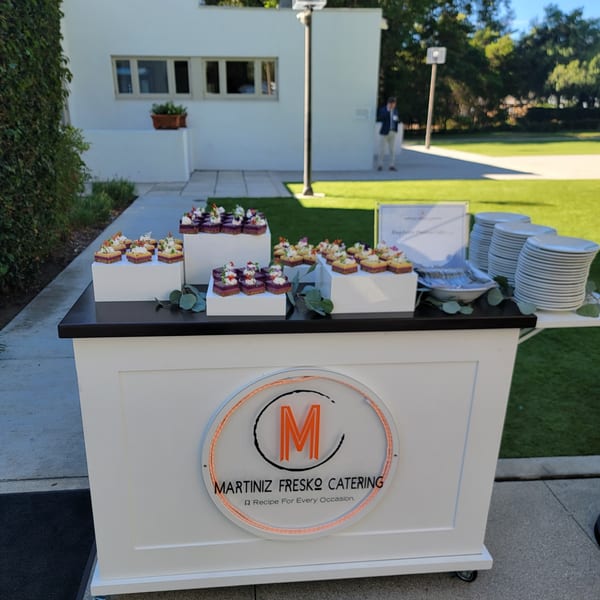 Martiniz Fresko Catering cart with small desserts, plates, and cutlery outdoors on a sunny day.