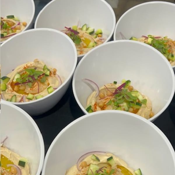 Several white bowls of hummus topped with chickpeas, cucumber, red onion, and herbs, ready for brunch.