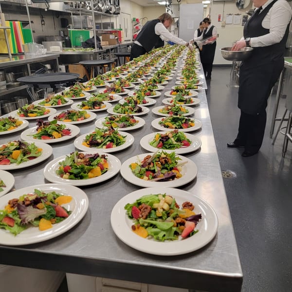 Chefs preparing numerous fresh fruit and nut salads on a long stainless steel kitchen counter.