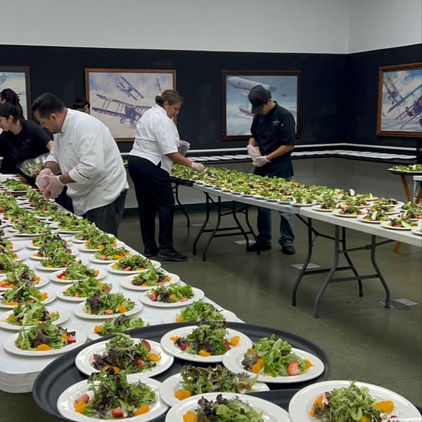 Chefs prepare numerous fresh salads with mixed greens, strawberries, and oranges on long tables.