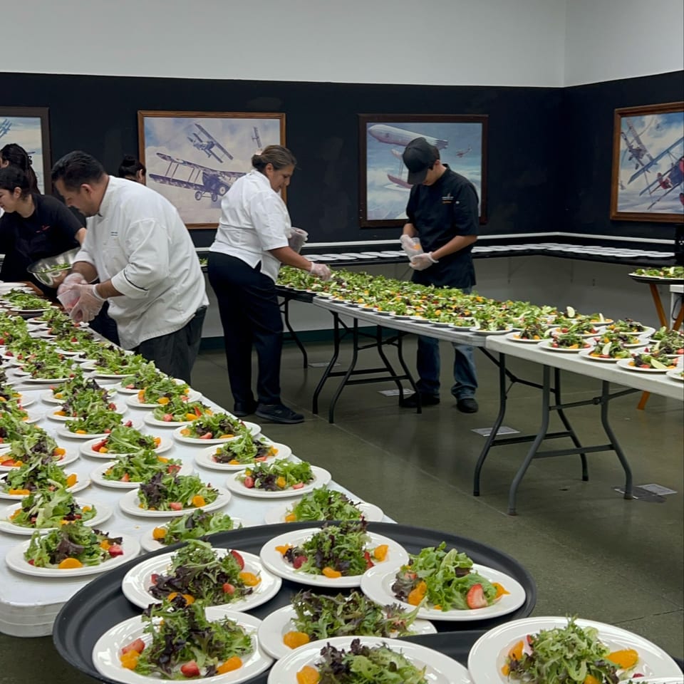 Chefs prepare numerous fresh salads with mixed greens, strawberries, and oranges on long tables.