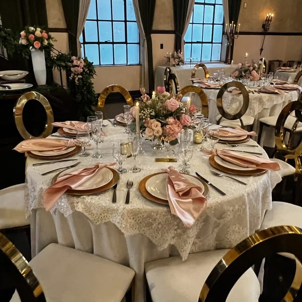 Elegant dining tables set with white lace tablecloths, gold-rimmed plates, pink napkins, and floral centerpieces.