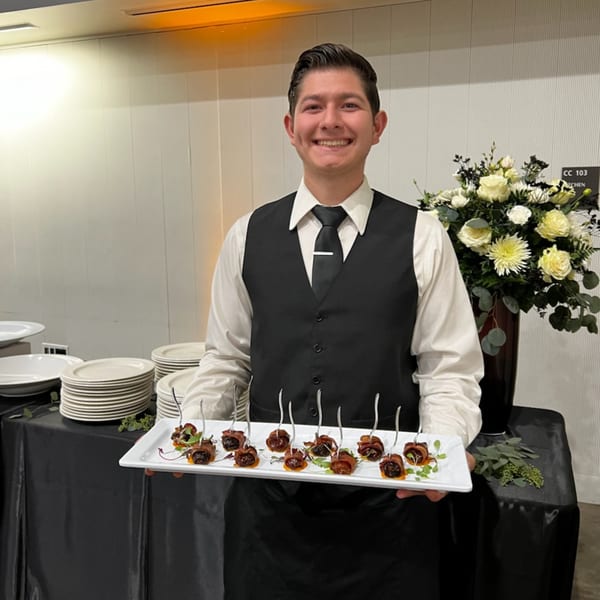 Smiling server in a black vest holding a tray of bacon-wrapped appetizers at Martiniz Fresko Kitchen.